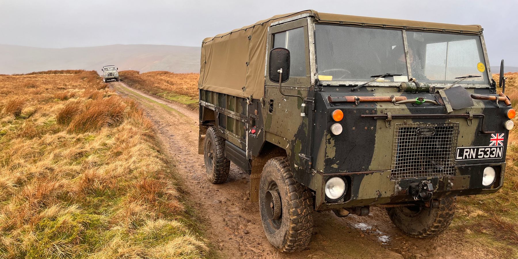 A camouflaged Land Rover 101 Forward Control on a muddy moorland track, with another vehicle further along the track